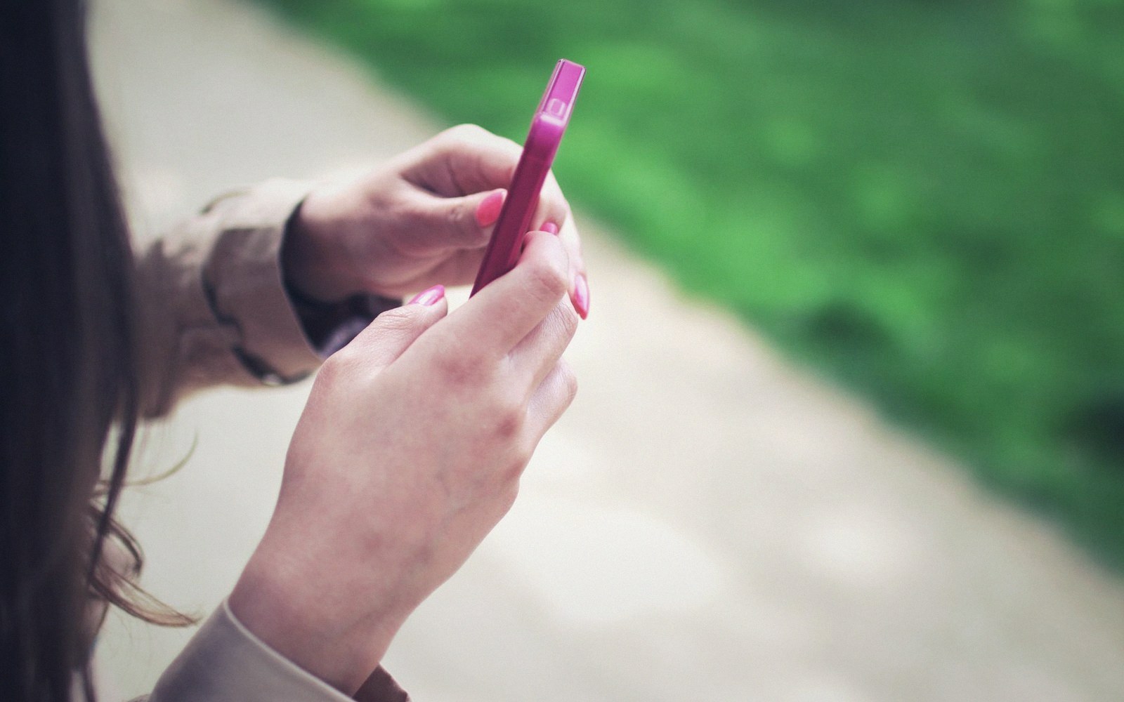 A woman holds a pink smartphone with both hands while standing on a path outdoors